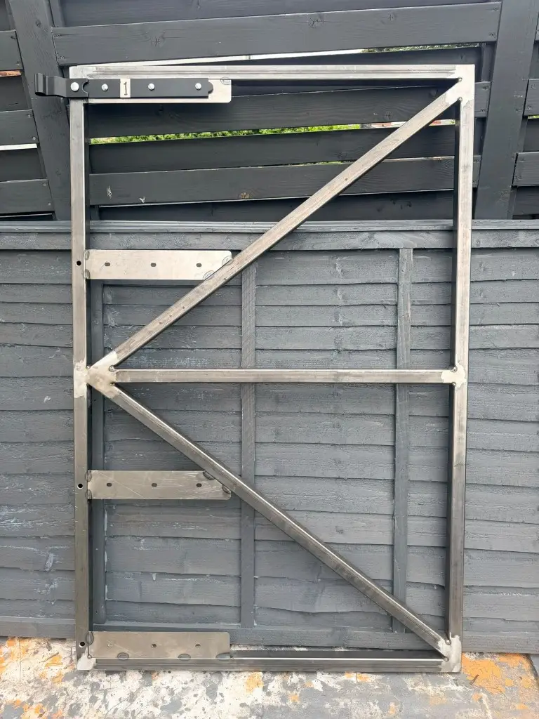 Custom-fabricated metal gate frame with diagonal bracing and mounting hardware, leaning against a dark wooden fence
