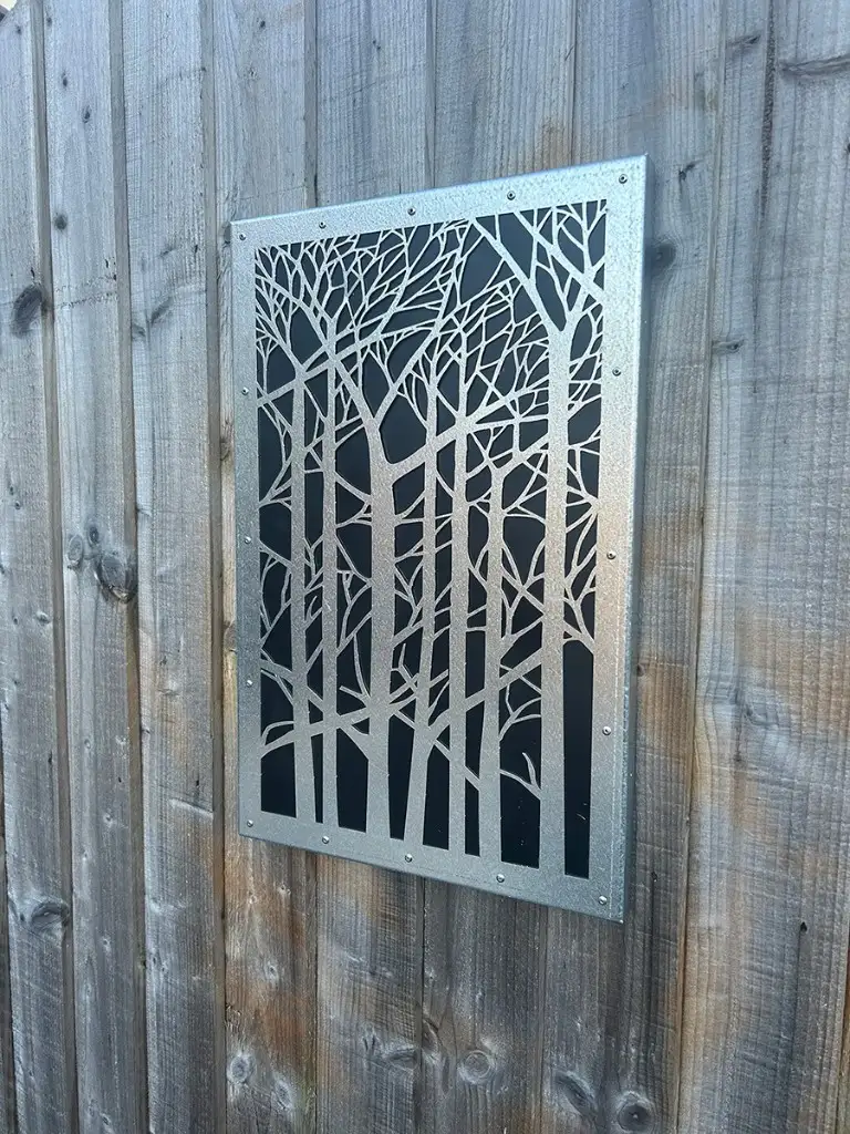 Silver laser-cut forest screen with black backing plate on a weathered fence