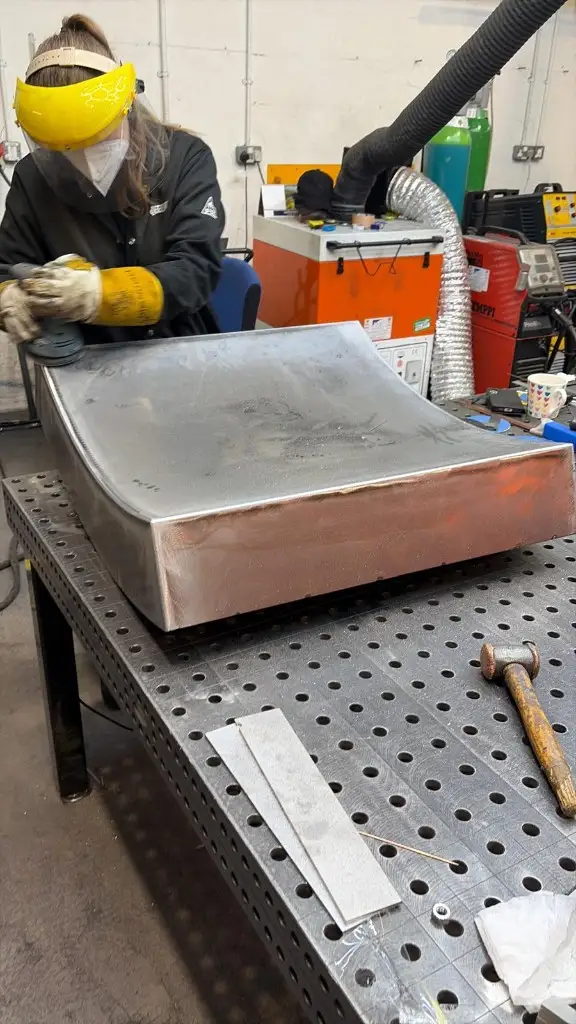 Fabricator in a face shield dressing the seams of a curved dining table base by hand