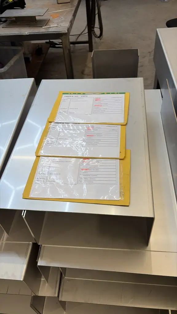 Stacked stainless magnet case outers with yellow job folders listing each manufacturing stage