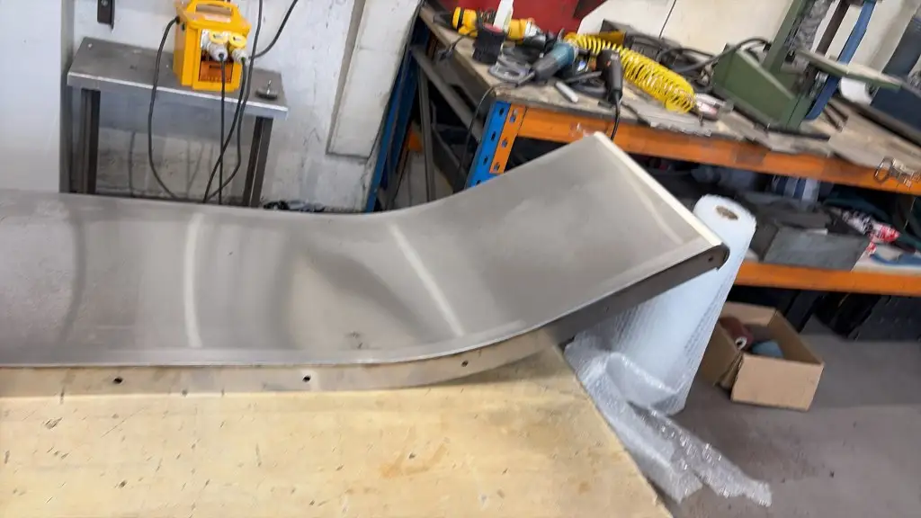 Rolled metal playground slide section with a satin finish and drilled side flange on a workshop bench